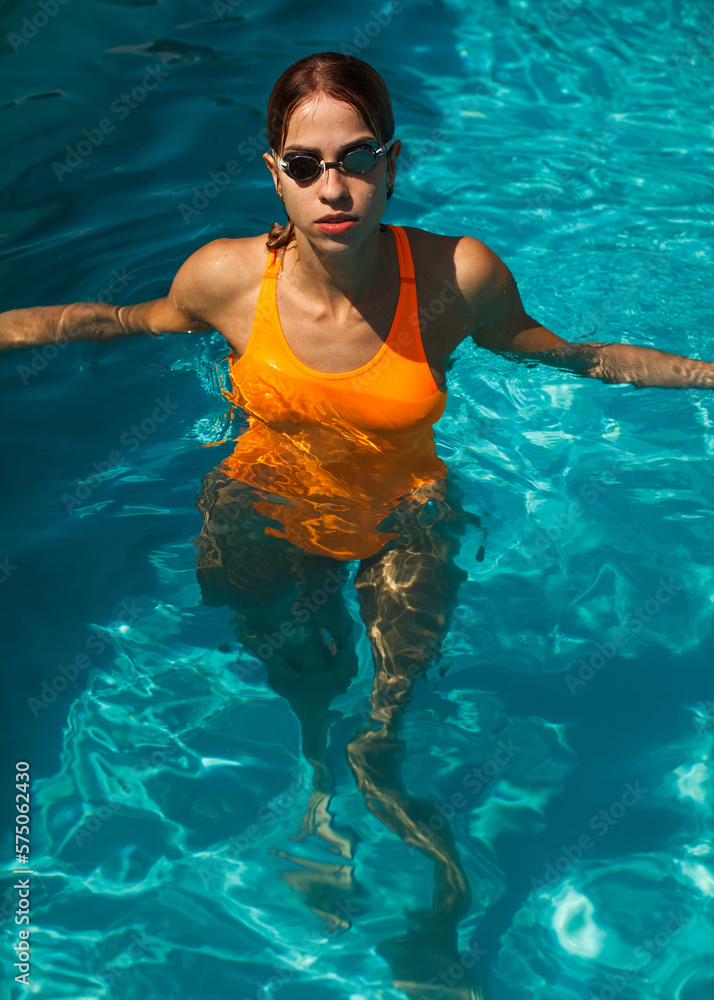 attractive athletic woman in orange swimsuit and goggles standing in open swimming pool during sunny day