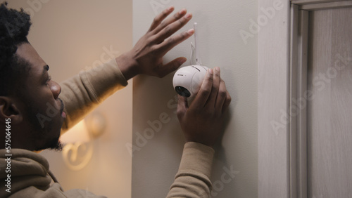 African American man puts security camera on wall fastening and connects it to system with cable. Man installs cameras in his apartment. Concept of CCTV cameras, tracking system, safety and privacy.