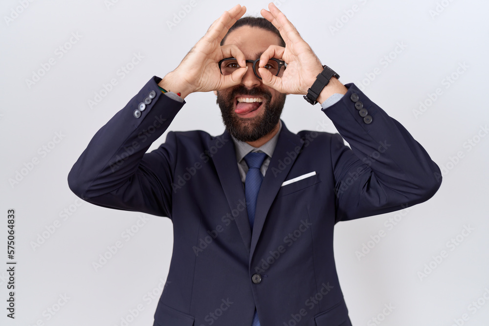 Hispanic man with beard wearing suit and tie doing ok gesture like binoculars sticking tongue out, eyes looking through fingers. crazy expression. Hispanic man with beard wearing suit and tie doing ok gesture like binoculars sticking tongue out, eyes looking through fingers. crazy expression.