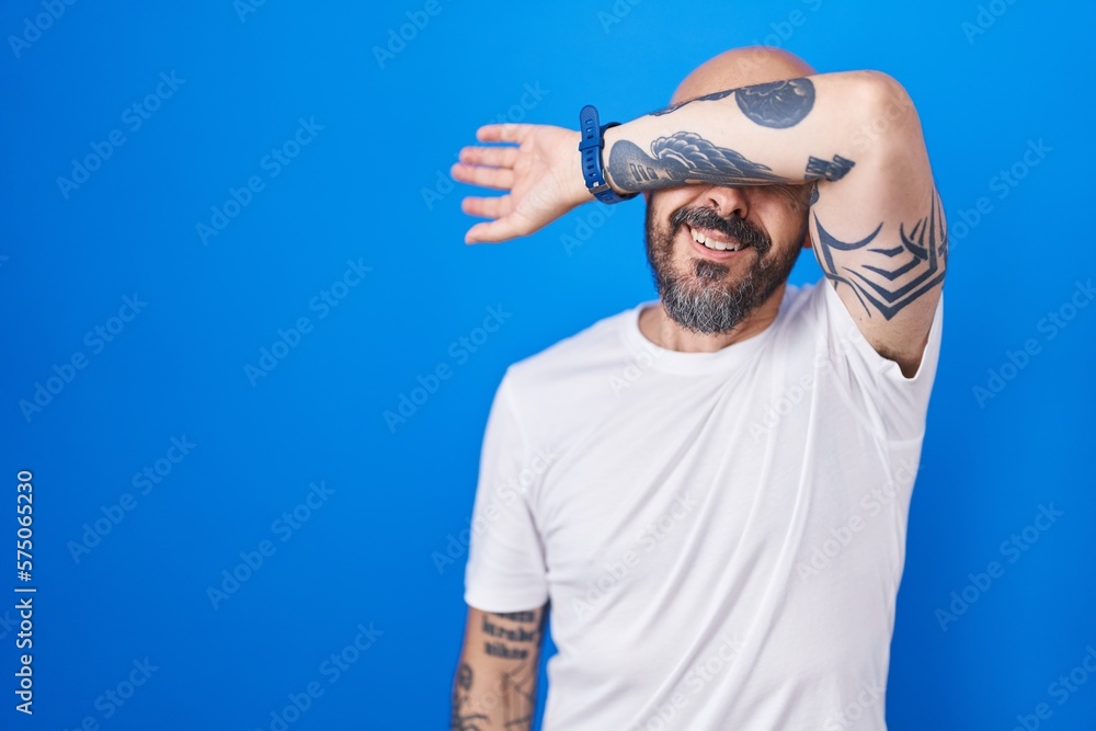 Naklejka premium Hispanic man with tattoos standing over blue background covering eyes with arm, looking serious and sad. sightless, hiding and rejection concept