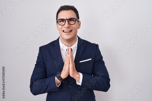 Young hispanic man wearing suit and tie begging and praying with hands together with hope expression on face very emotional and worried. begging.