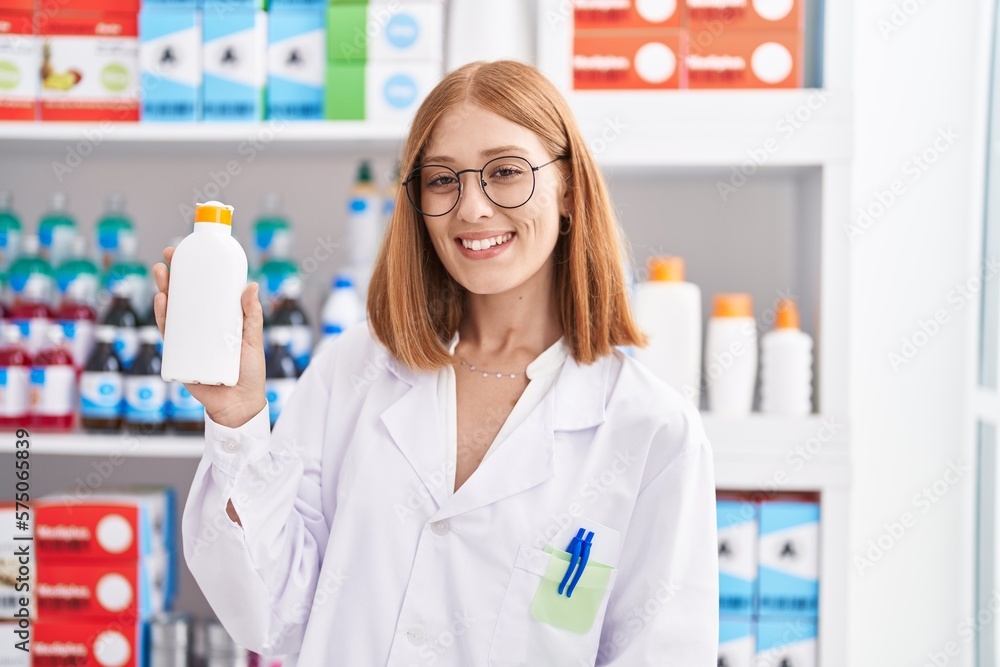 Young redhead woman working at pharmacy drugstore holding sun screen looking positive and happy standing and smiling with a confident smile showing teeth Young redhead woman working at pharmacy drugstore holding sun screen looking positive and happy standing and smiling with a confident smile showing teeth
