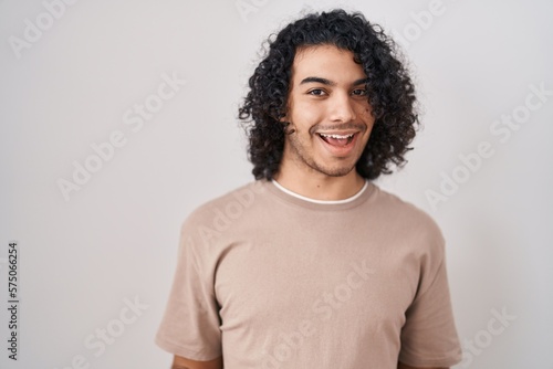 Hispanic man with curly hair standing over white background smiling cheerful with open arms as friendly welcome, positive and confident greetings