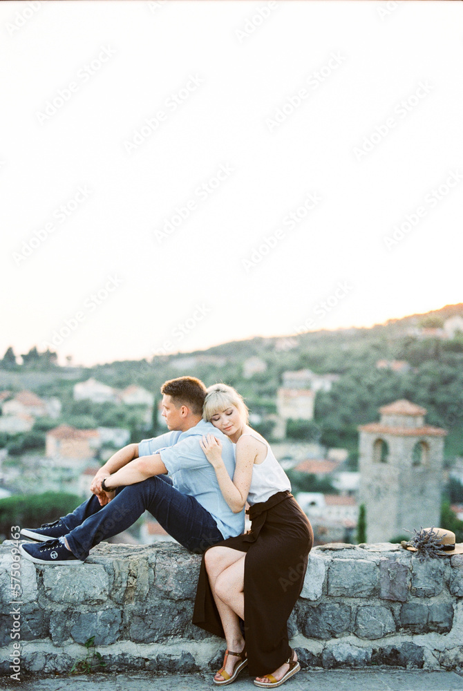 Woman hugs man from behind, resting her head on his shoulder, sitting on a stone fence