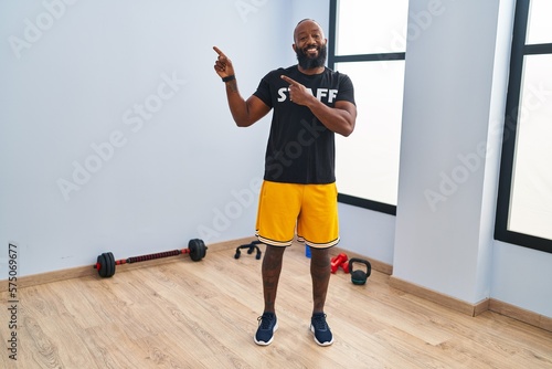 African american man working at fitness gym smiling and looking at the camera pointing with two hands and fingers to the side.