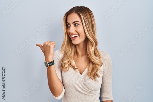 Young blonde woman standing over isolated background smiling with happy face looking and pointing to the side with thumb up.
