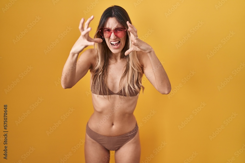 Young hispanic woman wearing bikini over yellow background shouting frustrated with rage, hands trying to strangle, yelling mad Young hispanic woman wearing bikini over yellow background shouting frustrated with rage, hands trying to strangle, yelling mad