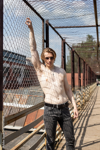 A guy with a bare torso and sunglasses. Portrait of a boy on the background of a metal grid. Look. Man in jeans. Muscles. Light and shadow