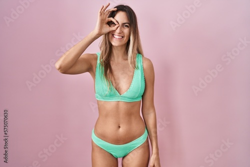 Young hispanic woman wearing bikini over pink background doing ok gesture with hand smiling, eye looking through fingers with happy face. © Krakenimages.com