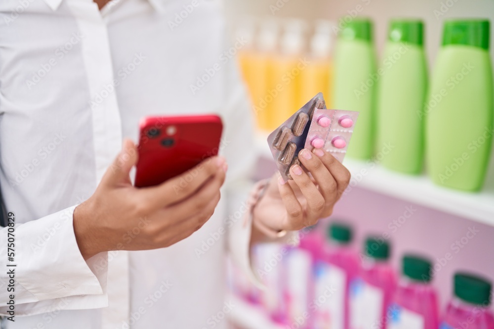 Young beautiful hispanic woman client using smartphone holding pills tablets at pharmacy