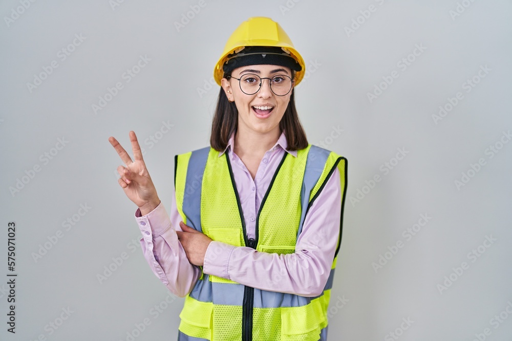 Hispanic girl wearing builder uniform and hardhat smiling with happy face winking at the camera doing victory sign. number two. Hispanic girl wearing builder uniform and hardhat smiling with happy face winking at the camera doing victory sign. number two.