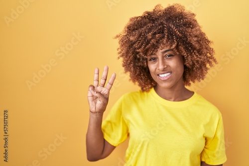 Young hispanic woman with curly hair standing over yellow background showing and pointing up with fingers number three while smiling confident and happy.