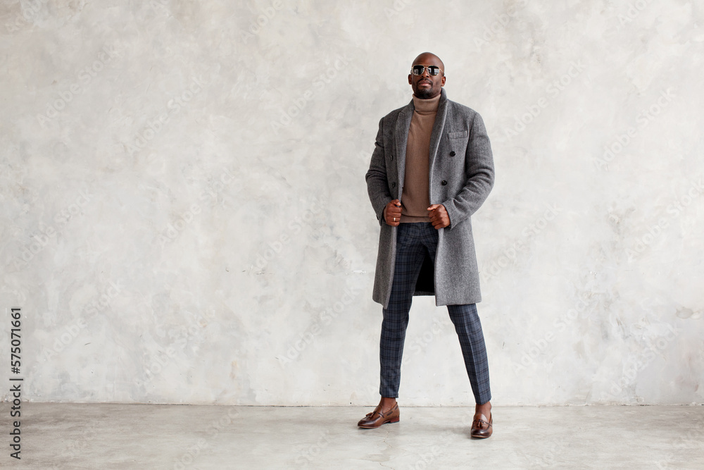 Handsome fashion man in turtleneck, gray coat, plaid trousers and leather loafer boots stands isolated on wall background, copy space. Fashionable male model