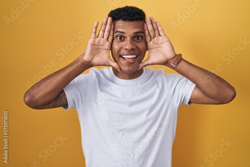 Young hispanic man standing over yellow background smiling cheerful playing peek a boo with hands showing face. surprised and exited © Krakenimages.com