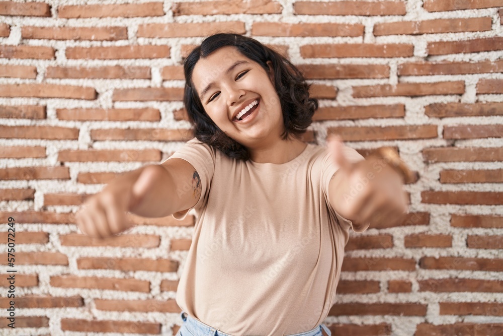 Young hispanic woman standing over bricks wall approving doing positive gesture with hand, thumbs up smiling and happy for success. winner gesture. Young hispanic woman standing over bricks wall approving doing positive gesture with hand, thumbs up smiling and happy for success. winner gesture.