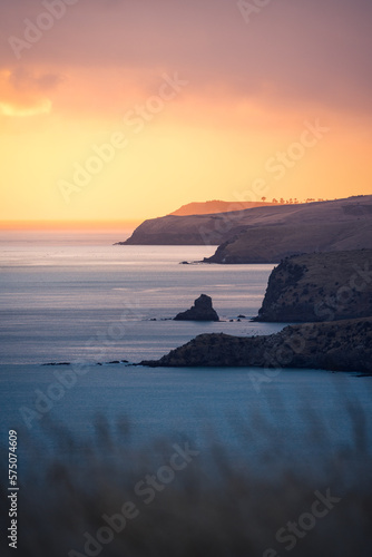 Steile Küste im orangem Himmel in der Dämmerung zum Sonnenuntergang mit Gras im Vordergrund in Neuseeland.