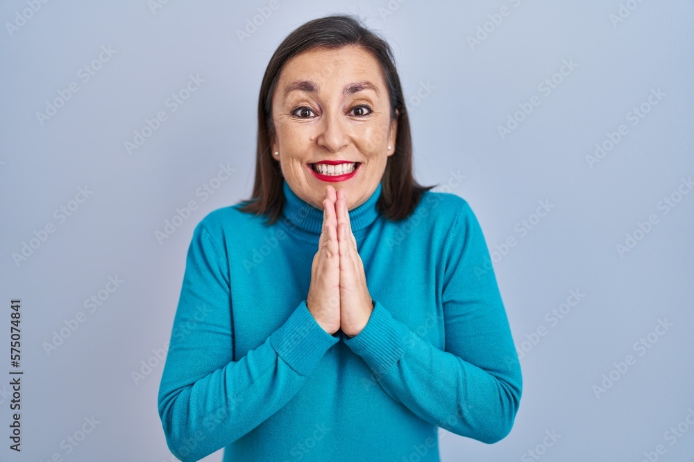 Middle age hispanic woman standing over isolated background praying with hands together asking for forgiveness smiling confident.