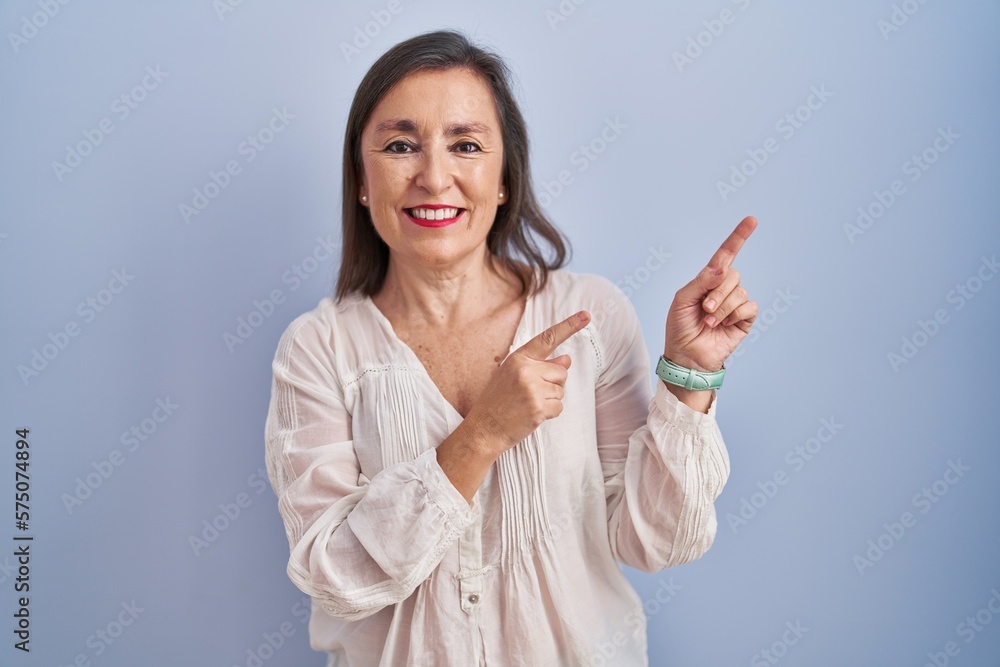 Middle age hispanic woman standing over blue background smiling and looking at the camera pointing with two hands and fingers to the side. Middle age hispanic woman standing over blue background smiling and looking at the camera pointing with two hands and fingers to the side.