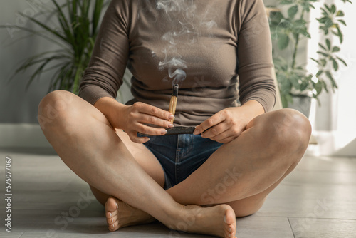 a faceless middle-aged woman in a beige turtleneck holds a wooden palo santo wand with a magical stream of smoke before meditation and yoga, sitting on the floor of the house