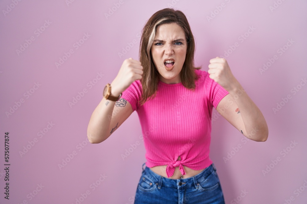 Blonde caucasian woman standing over pink background angry and mad raising fists frustrated and furious while shouting with anger. rage and aggressive concept. Blonde caucasian woman standing over pink background angry and mad raising fists frustrated and furious while shouting with anger. rage and aggressive concept.