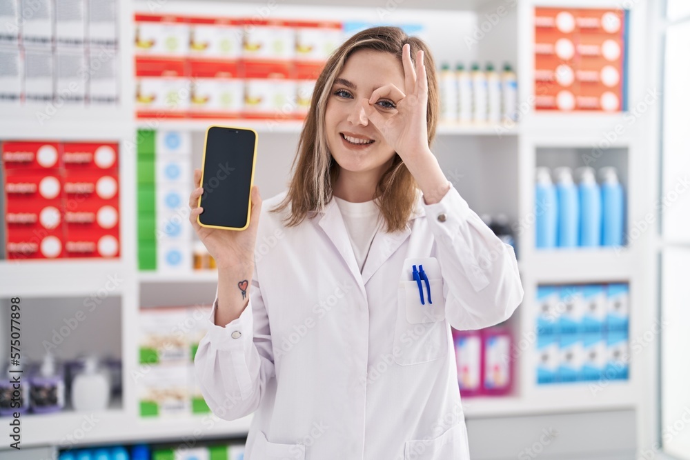 Blonde caucasian woman working at pharmacy drugstore showing smartphone screen smiling happy doing ok sign with hand on eye looking through fingers
