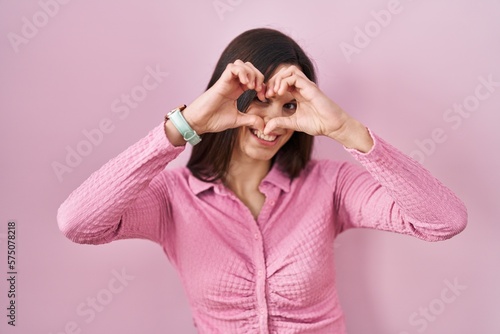 Young hispanic woman standing over pink background doing heart shape with hand and fingers smiling looking through sign © Krakenimages.com