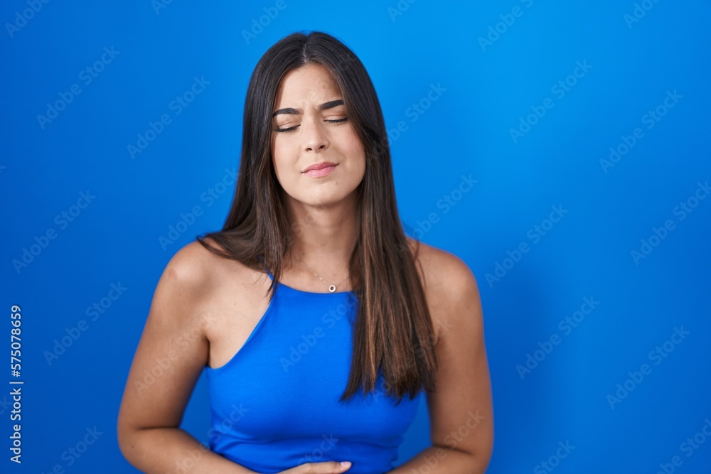 Hispanic woman standing over blue background with hand on stomach because indigestion, painful illness feeling unwell. ache concept. Hispanic woman standing over blue background with hand on stomach because indigestion, painful illness feeling unwell. ache concept.