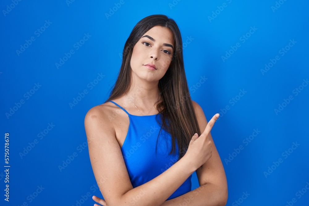 Hispanic woman standing over blue background pointing with hand finger to the side showing advertisement, serious and calm face Hispanic woman standing over blue background pointing with hand finger to the side showing advertisement, serious and calm face