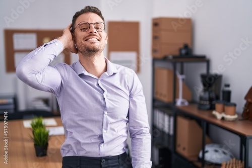 Young hispanic man at the office smiling confident touching hair with hand up gesture, posing attractive and fashionable