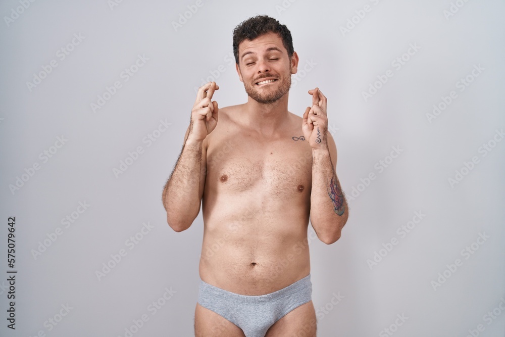 Young hispanic man standing shirtless wearing underware gesturing finger crossed smiling with hope and eyes closed. luck and superstitious concept.