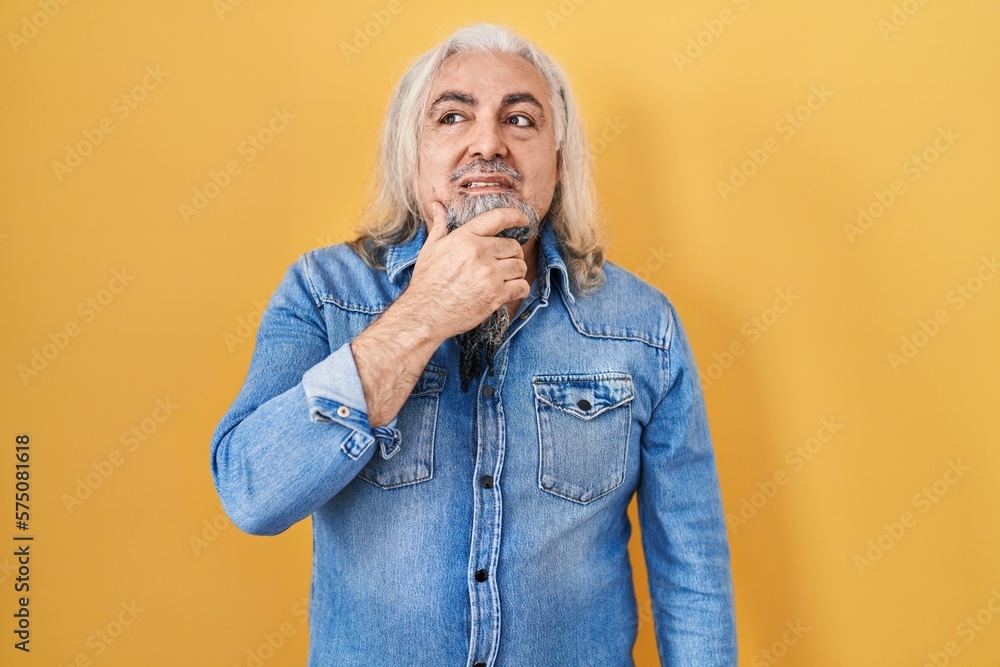 Middle age man with grey hair standing over yellow background thinking worried about a question, concerned and nervous with hand on chin Middle age man with grey hair standing over yellow background thinking worried about a question, concerned and nervous with hand on chin