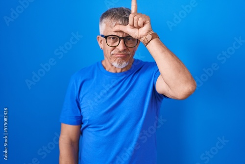 Hispanic man with grey hair standing over blue background making fun of people with fingers on forehead doing loser gesture mocking and insulting.