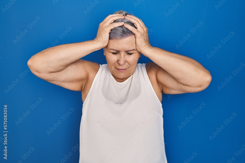 Middle age caucasian woman standing over blue background suffering from headache desperate and stressed because pain and migraine. hands on head. Middle age caucasian woman standing over blue background suffering from headache desperate and stressed because pain and migraine. hands on head.