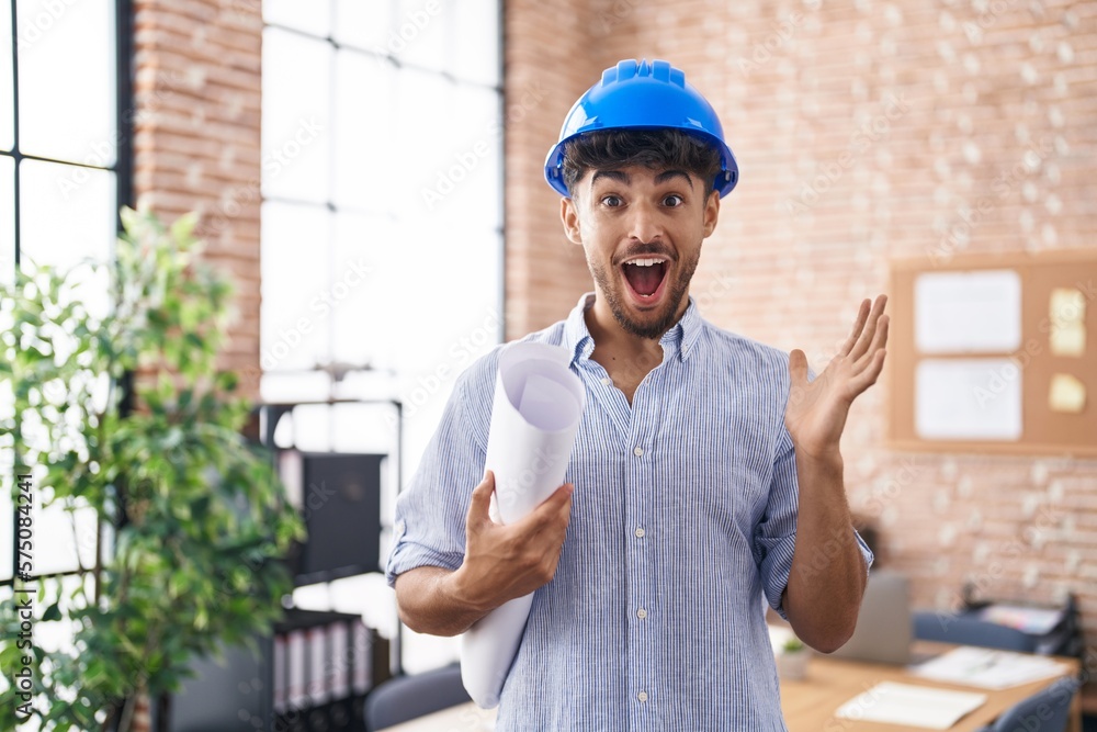 Arab man with beard wearing architect hardhat at construction office celebrating victory with happy smile and winner expression with raised hands Arab man with beard wearing architect hardhat at construction office celebrating victory with happy smile and winner expression with raised hands