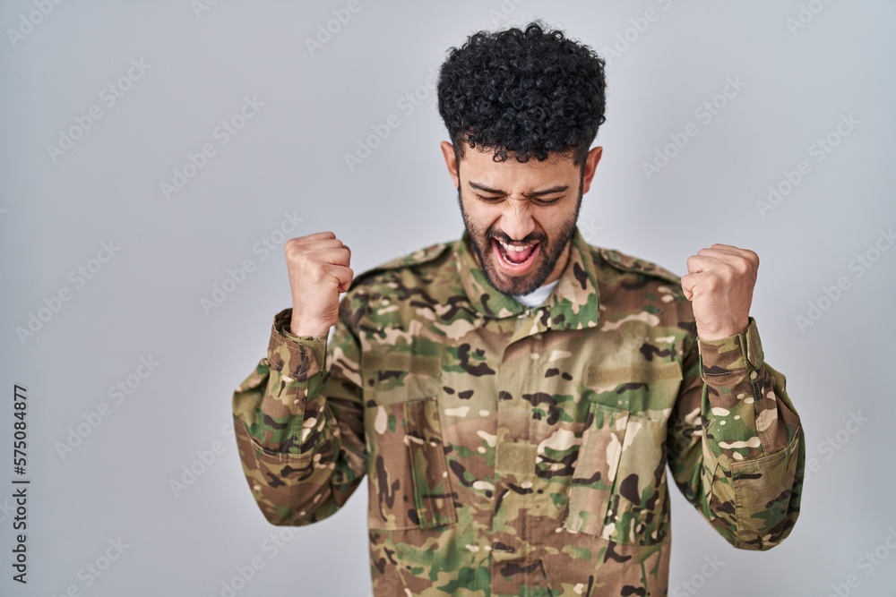 Arab man wearing camouflage army uniform celebrating mad and crazy for success with arms raised and closed eyes screaming excited. winner concept