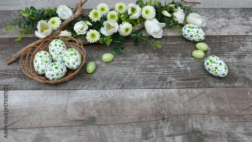Ein Korb mit Ostereiern umrahmt von Blumen in der Farbe weiß und grün auf altem Holz Hintergrund mit Platz für Text.