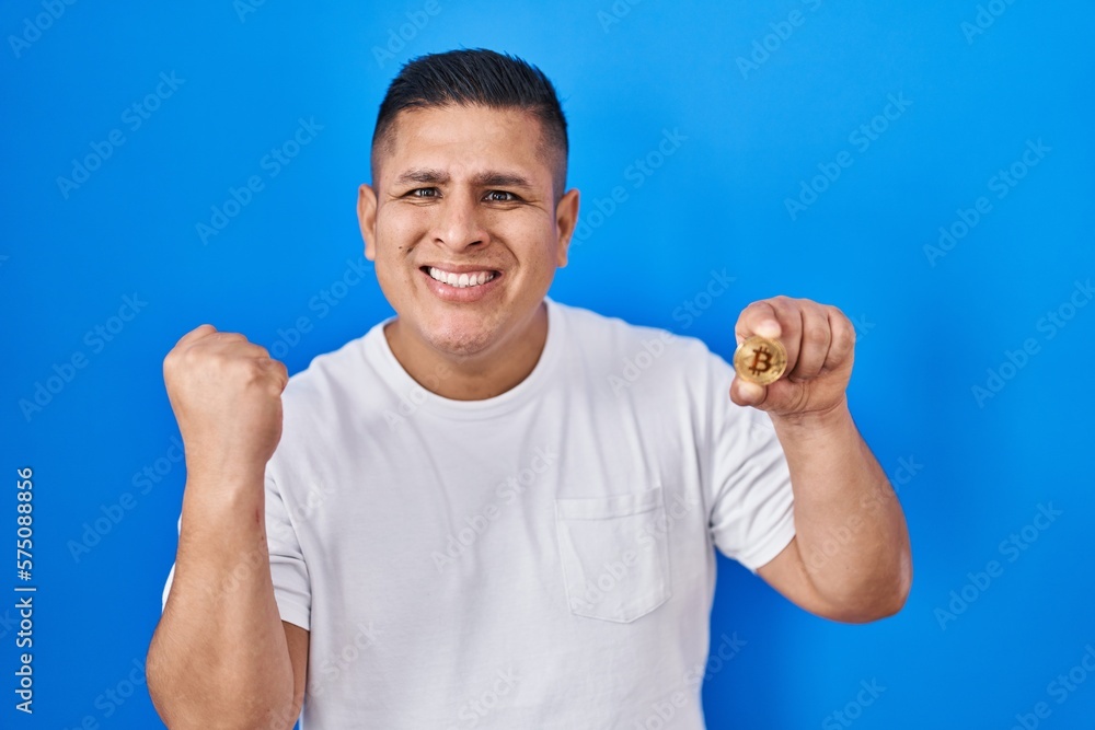 Fototapeta premium Hispanic young man holding virtual currency bitcoin screaming proud, celebrating victory and success very excited with raised arms