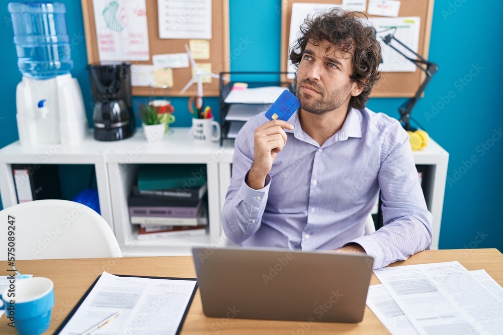 Young hispanic man business worker using laptop and credit card with doubt expression at office Young hispanic man business worker using laptop and credit card with doubt expression at office