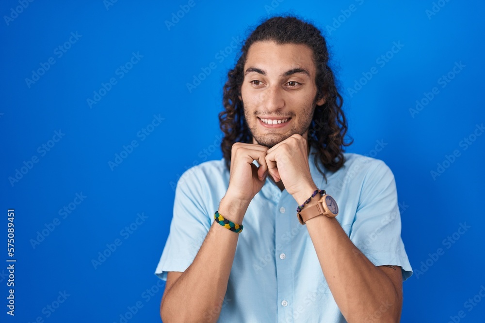 Young hispanic man standing over blue background laughing nervous and excited with hands on chin looking to the side Young hispanic man standing over blue background laughing nervous and excited with hands on chin looking to the side