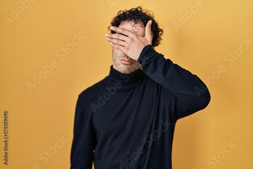 Hispanic man standing over yellow background covering eyes with hand, looking serious and sad. sightless, hiding and rejection concept