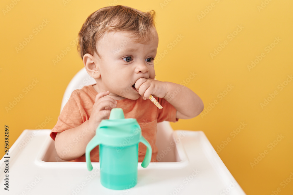 Adorable blond toddler sitting on highchair eating snack over isolated yellow background Adorable blond toddler sitting on highchair eating snack over isolated yellow background