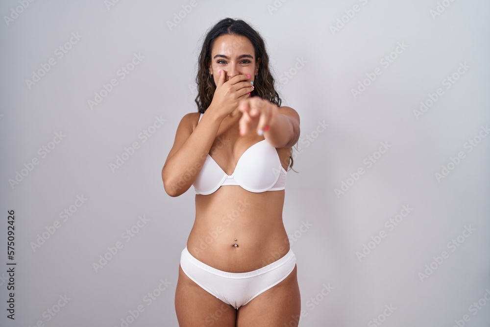 Fototapeta premium Young hispanic woman wearing white lingerie laughing at you, pointing finger to the camera with hand over mouth, shame expression