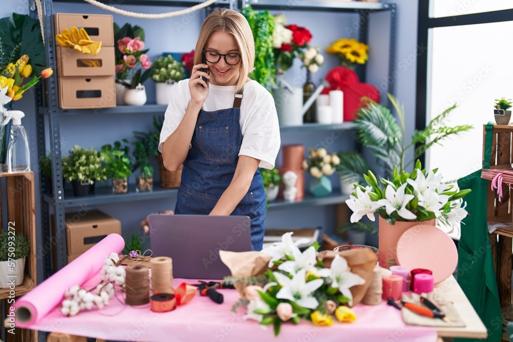 Young blonde woman florist using laptop talking on smartphone at florist shop Young blonde woman florist using laptop talking on smartphone at florist shop