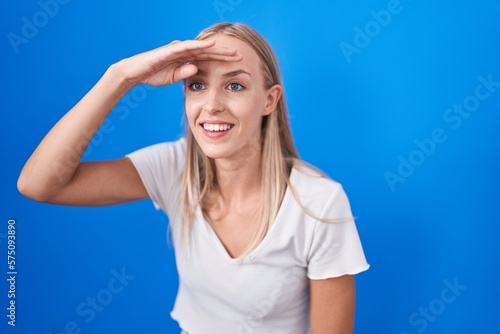 Young caucasian woman standing over blue background very happy and smiling looking far away with hand over head. searching concept.