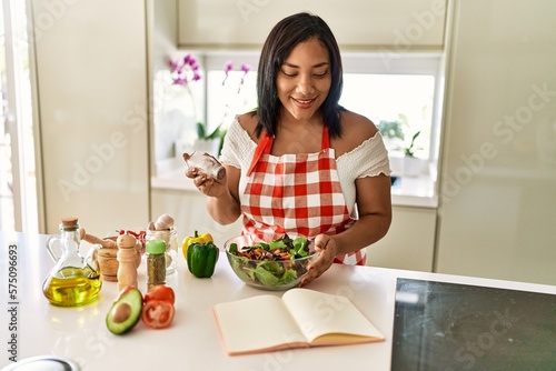 Hispanic brunette woman seasoning green salad at the kitchen