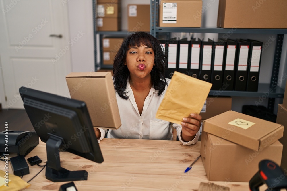 Hispanic woman working at small business ecommerce holding packages puffing cheeks with funny face. mouth inflated with air, catching air. Hispanic woman working at small business ecommerce holding packages puffing cheeks with funny face. mouth inflated with air, catching air.