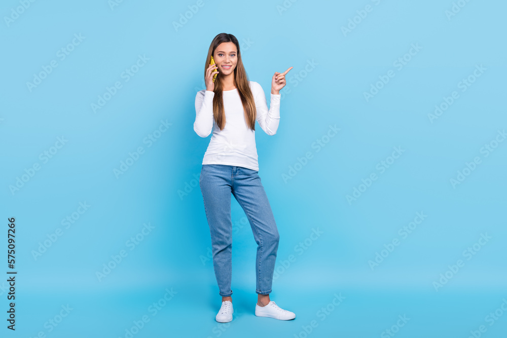 Full length photo of shiny cute woman wear white shirt talking gadget pointing empty space isolated blue color background Full length photo of shiny cute woman wear white shirt talking gadget pointing empty space isolated blue color background