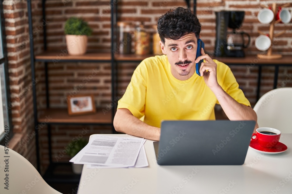 Young caucasian man using laptop talking on smartphone at home Young caucasian man using laptop talking on smartphone at home