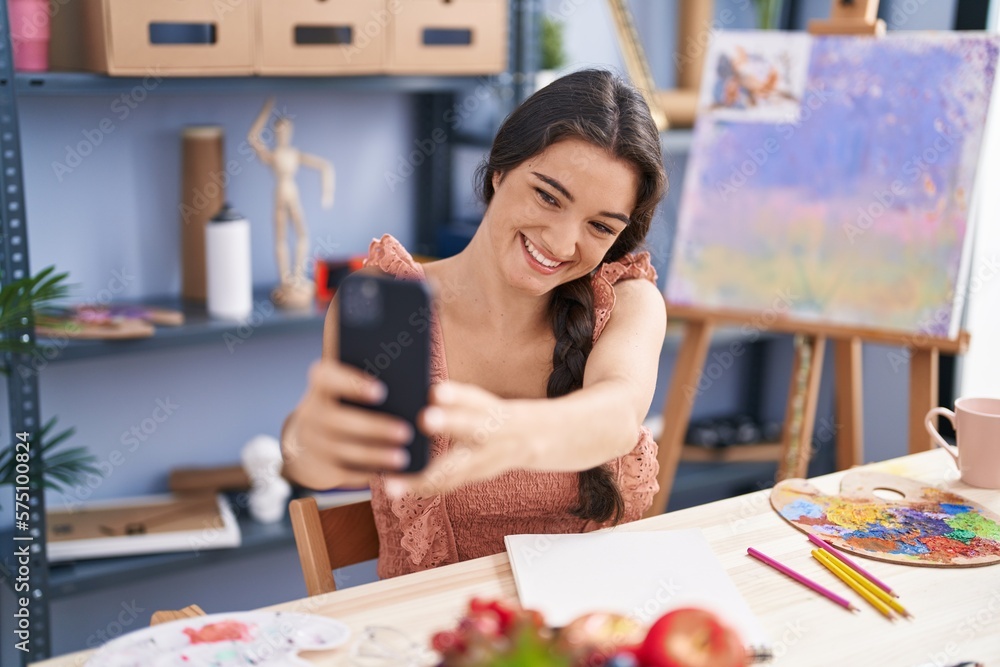 Fototapeta premium Young hispanic woman artist drawing on notebook make selfie by smartphone at art studio