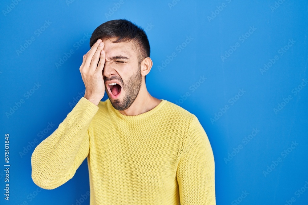 Hispanic man standing over blue background yawning tired covering half face, eye and mouth with hand. face hurts in pain. Hispanic man standing over blue background yawning tired covering half face, eye and mouth with hand. face hurts in pain.